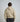 A man viewed from behind wearing a light beige pullover hoodie and dark pants, standing against a plain light gray studio background.
