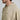 Close-up of a man wearing a light beige pullover hoodie with a small black stylized logo on the left chest, against a plain light gray background.